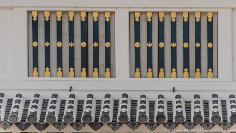 Black barred windows with gold fittings above gray roof tiles on a white castle wall.
