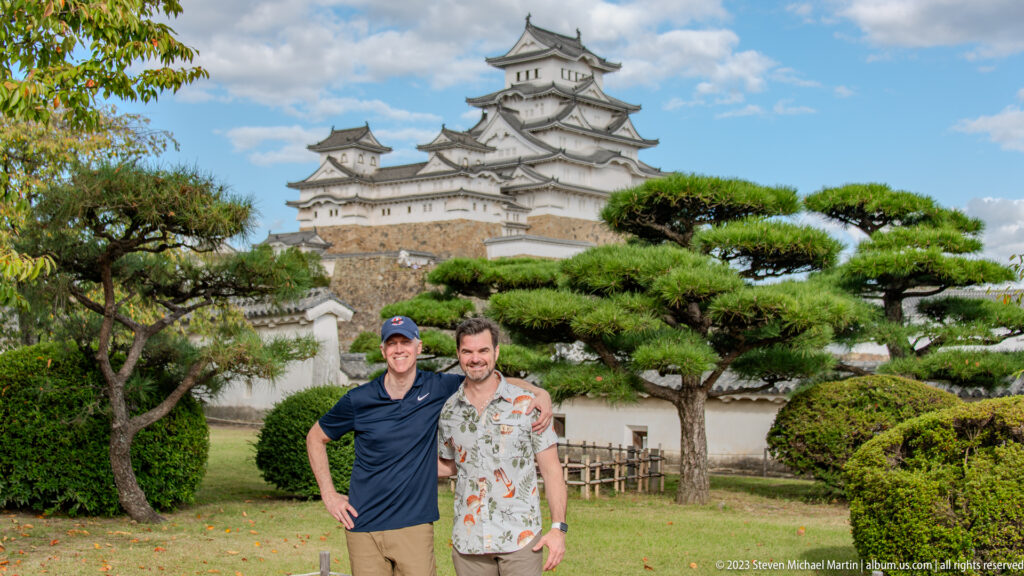 Two visitors posing among sculpted pine trees with Himeji Castle behind them.