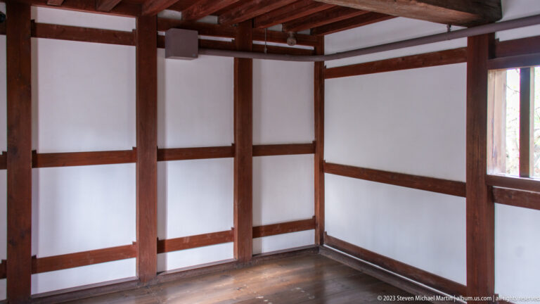 Empty wooden room with exposed dark beams, white plaster walls, and a rectangular window.