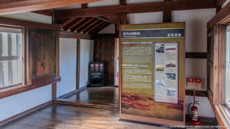 Tall historical interpretation panel standing in a timber corridor with exposed beams, wooden shutters, polished floors, and white plaster walls at Himeji Castle.