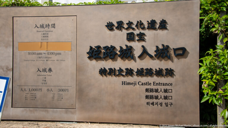 Stone Himeji Castle entrance sign beside a panel listing opening hours and admission fees.