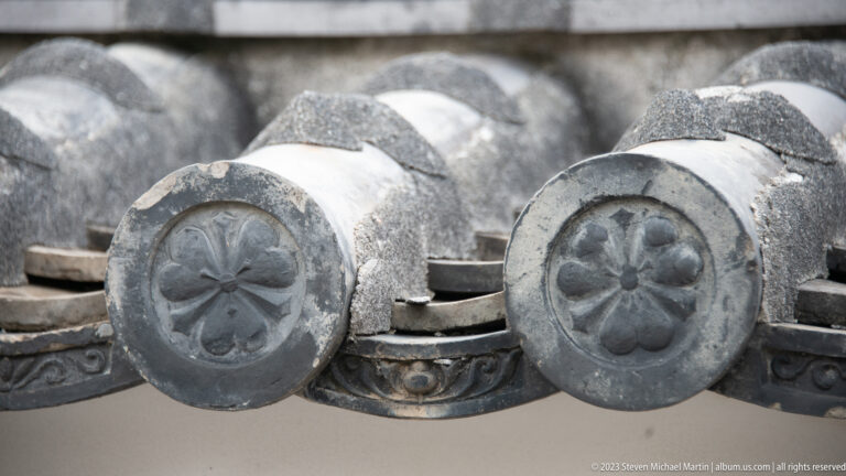 Close view of two gray circular eave-end tiles with matching floral crests above a molded decorative roof-tile band at Himeji Castle.