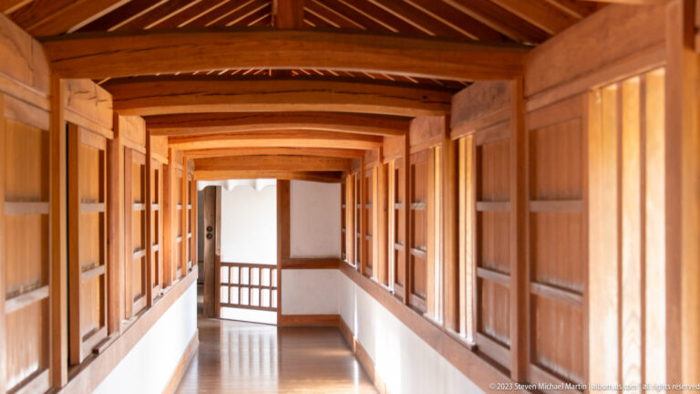 Long sunlit wooden gallery with open shutters, exposed rafters, repeating timber posts, white lower walls, and a polished plank floor inside Himeji Castle.