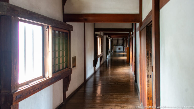 Long dim corridor inside Himeji Castle with open wooden shutters on the left, dark timber framing, white plaster walls, and a reflective plank floor.