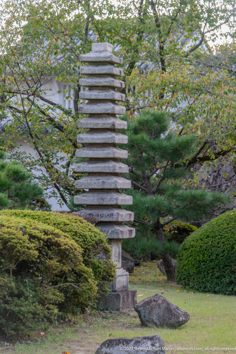 Tall multi-tiered stone pagoda standing among moss, clipped shrubs, pines, rocks, and leafy trees in a Japanese garden near Himeji Castle.