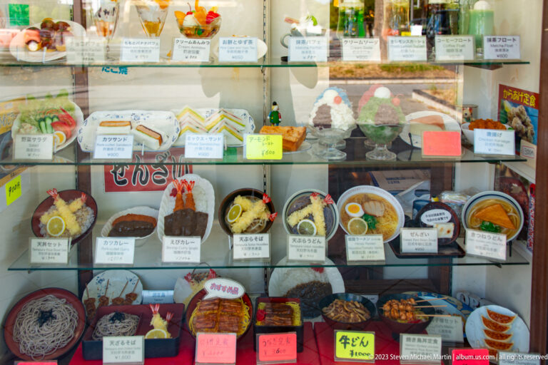 Glass restaurant case filled with plastic models of Japanese meals, including noodle bowls, rice dishes, curry, fried foods, desserts, drinks, and labeled set menus near Himeji Castle.