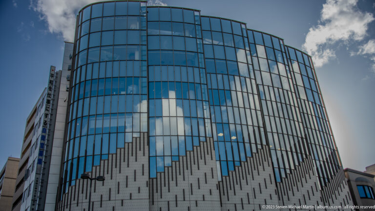 Curved modern building with a blue reflective glass facade, pale lower cladding, narrow vertical openings, and clouds mirrored across the upper stories in central Himeji.