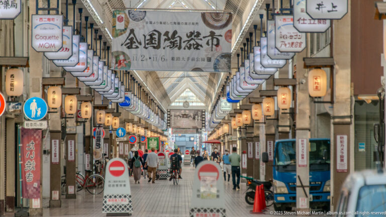 Long covered shopping arcade in Himeji with hanging lanterns, shop signs, banners, pedestrians, bicycles, and a translucent roof receding into the distance.