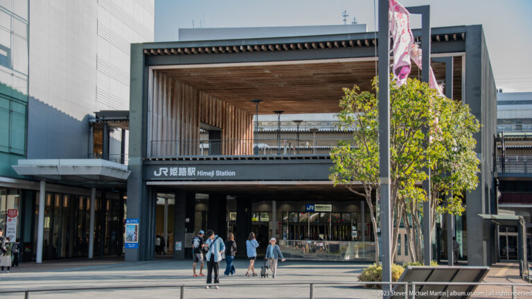 Main entrance to JR Himeji Station with bilingual station signage, a large rectangular timber-lined canopy, glass doors, trees, and pedestrians in the plaza.