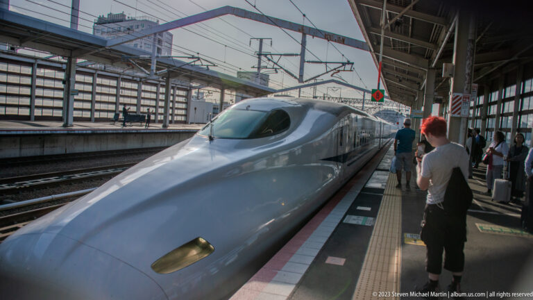 White Shinkansen high-speed train with a long streamlined nose standing at a station platform under overhead wires, with passengers nearby.