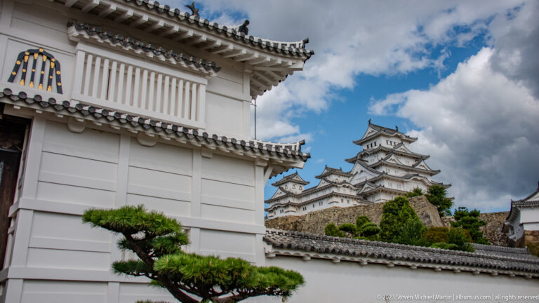 Himeji Castle’s main keep framed by a white gatehouse wall, tiled roofs, and a pruned pine.