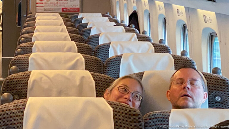 Interior of a Shinkansen carriage with rows of high-backed seats and two travelers leaning into the foreground.