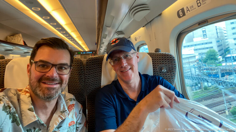 Two smiling travel companions seated together beside a window inside a Shinkansen train.