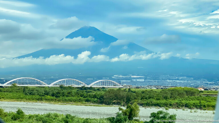 Mount Fuji rising through low clouds above a broad river plain, with a white arched bridge, green fields, and blue sky in the foreground.