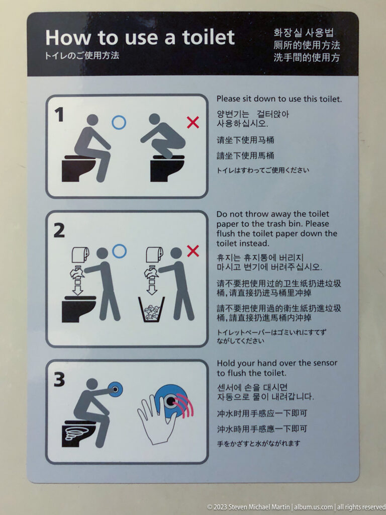 Multilingual restroom instruction sign with three pictograms showing correct seated toilet use, disposal of toilet paper in the bowl, and sensor-activated flushing.