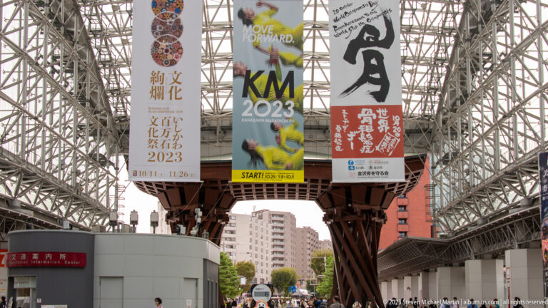 Glass lattice canopy, event banners, and part of the timber Tsuzumi-mon Gate above the Kanazawa Station approach.