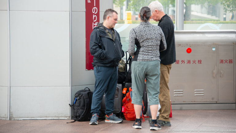 Three travelers talking together beside several backpacks and travel bags outside Kanazawa Station.