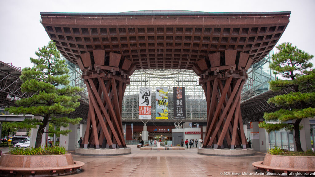 Centered frontal view of Kanazawa Station’s Tsuzumi-mon Gate on a wet forecourt.