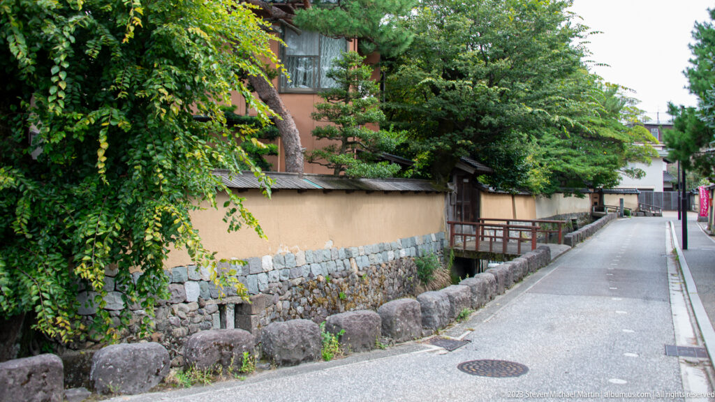 Narrow lane beside a stone-lined water channel, plaster wall, greenery, and small wooden bridge.