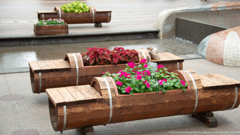 Two log-shaped wooden planters filled with pink and red flowers on a paved plaza.