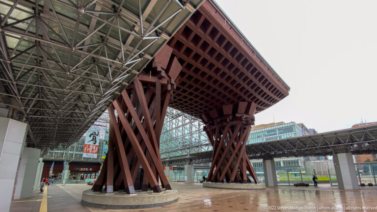Oblique side view of Tsuzumi-mon Gate beneath steel station canopies on wet pavement.