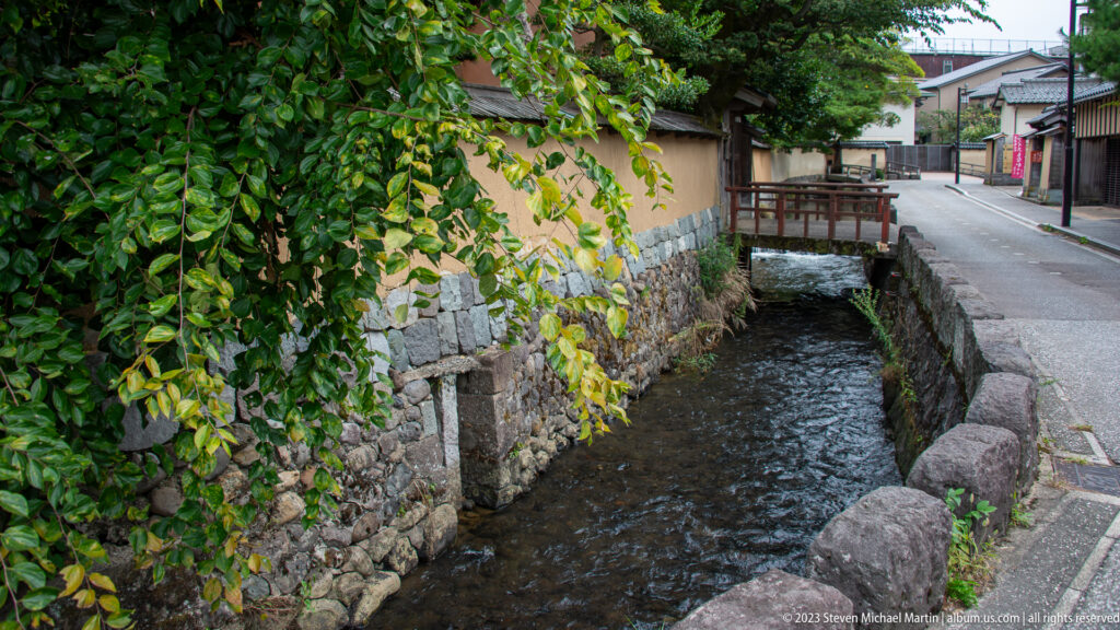 Stone-lined canal with overhanging vines, plaster wall, small bridge, and residential lane.