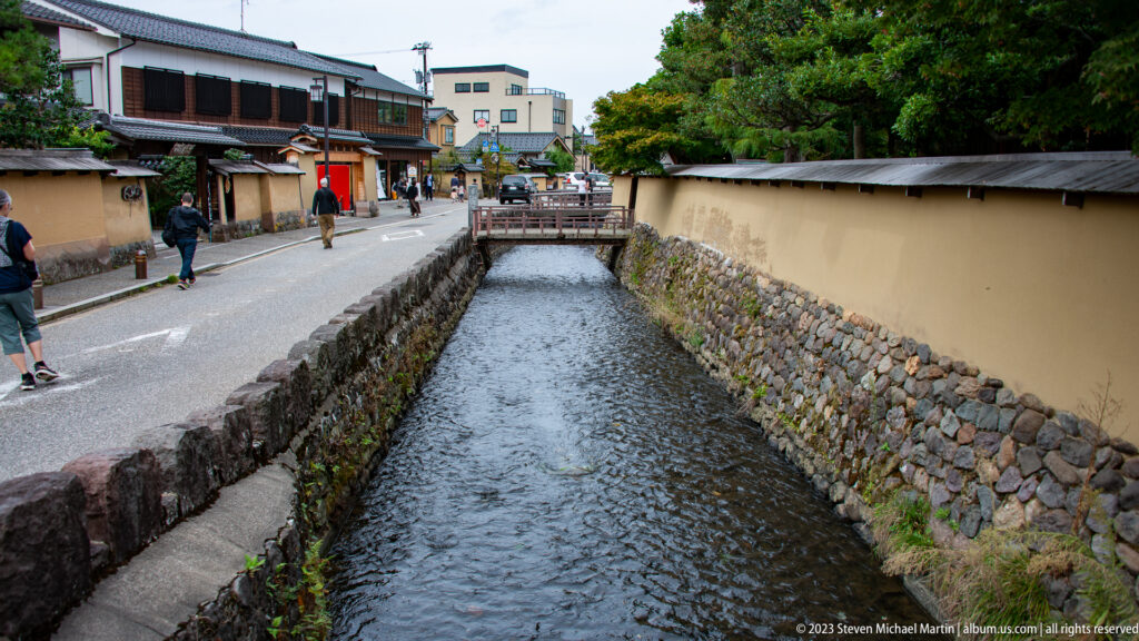 Straight canal between plaster walls and stone banks, crossed by a wooden bridge with pedestrians nearby.