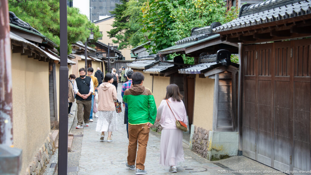 Visitors walking along a narrow lane between plaster walls, timber gates, tiled eaves, and greenery.