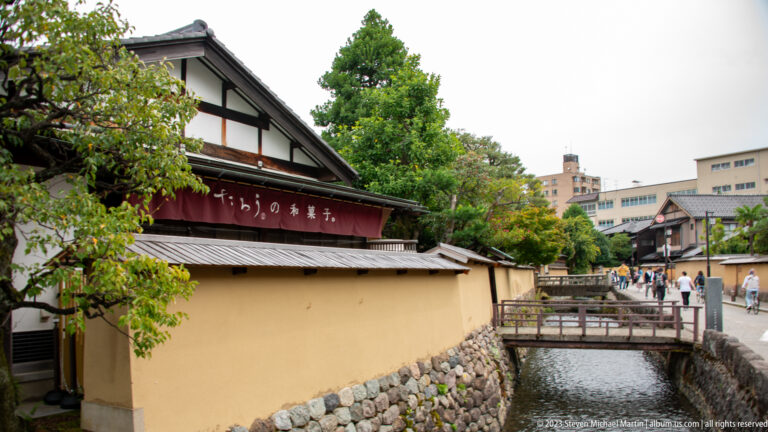 Traditional canal-side building with plaster wall, stone base, Japanese sign, tiled eaves, and bridge.