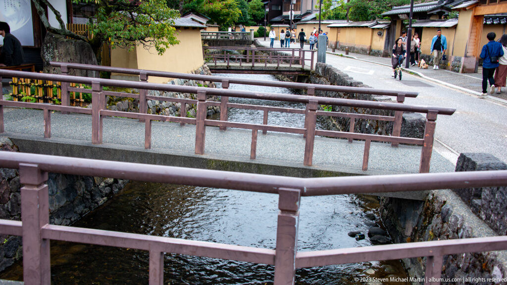Multiple pedestrian bridges crossing a narrow canal with stone banks, walls, and visitors beyond.