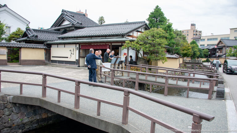 Visitors near a white-and-timber traditional building reached by a bridge beside a canal.