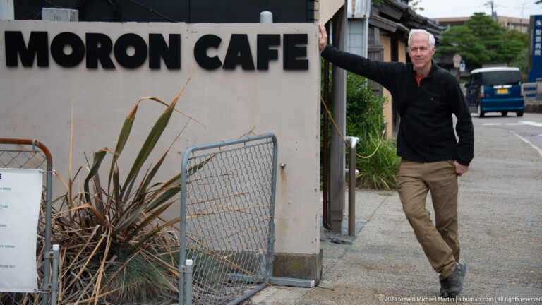Visitor raising one arm beside a cropped MACHI CAFE sign on a quiet Kanazawa street.