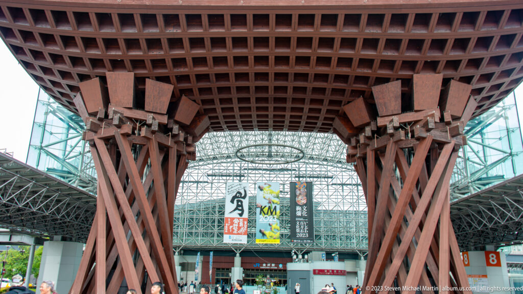 Upward view beneath the Tsuzumi-mon Gate, with bundled timber columns and the glass station dome behind.