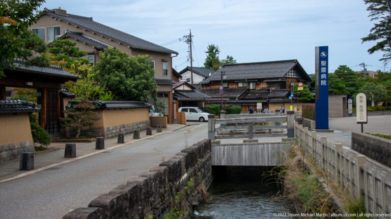 Stone-lined canal, bridge, tiled-roof houses, garden walls, and a tall blue entrance sign.