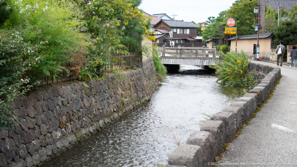 Narrow flowing canal between vegetated stone banks, with a small bridge and traditional buildings ahead.