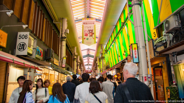 Crowded Omicho Market arcade with striped roof panels, numbered stalls, lanterns, and shoppers.