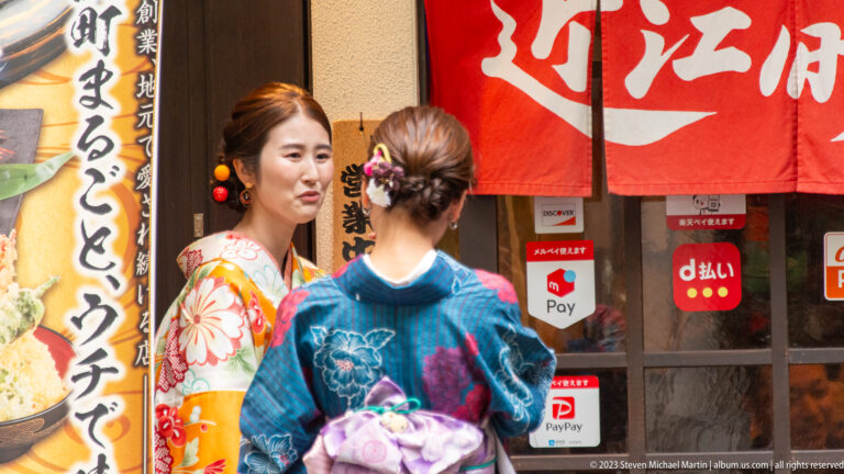 Two women in colorful kimono and obi outside a restaurant with red curtains and Japanese food signs.