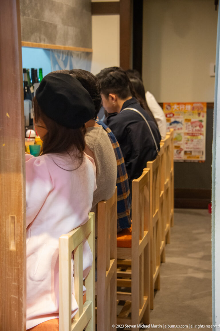 Diners seated closely on wooden chairs at a narrow counter inside a small market restaurant.