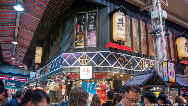 Busy Omicho Market junction with wooden shopfronts, lanterns, numbered signs, banners, and shoppers.