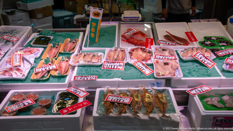 Chilled market counter filled with packaged fish and seafood marked by handwritten Japanese price labels.