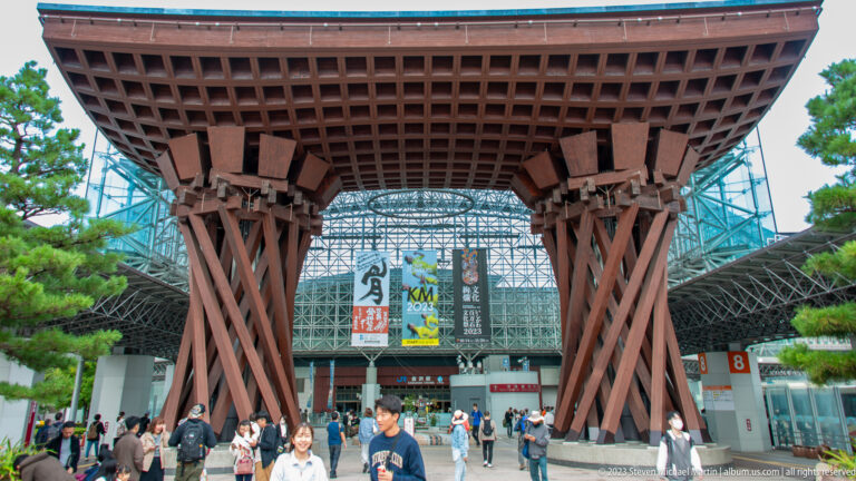 Full front view of the Tsuzumi-mon Gate with pedestrians, pines, and the glass station canopy beyond.