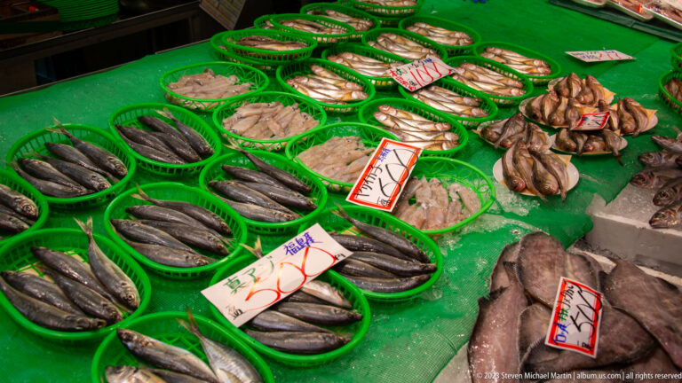 Assorted whole fish and shellfish arranged in green baskets with handwritten Japanese price cards.