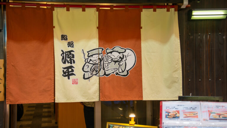 Orange and cream noren curtain with Japanese calligraphy and a cartoon emblem above a restaurant entrance.