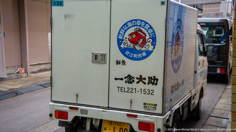 Compact delivery truck with Japanese lettering and a circular fish logo parked in a narrow lane.