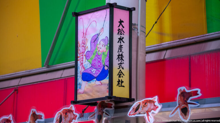 Illuminated vertical sign with Japanese lettering and a painted seafood figure against a colorful storefront.