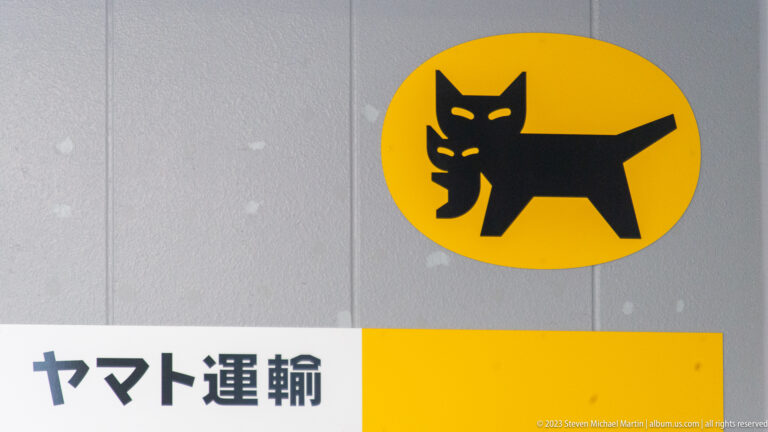 Black cat carrying a kitten inside a yellow oval above Japanese Yamato Transport lettering.