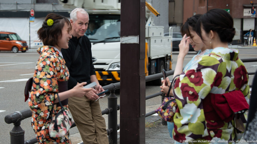 Two women in colorful floral kimono talking on a city street with a visitor behind them.