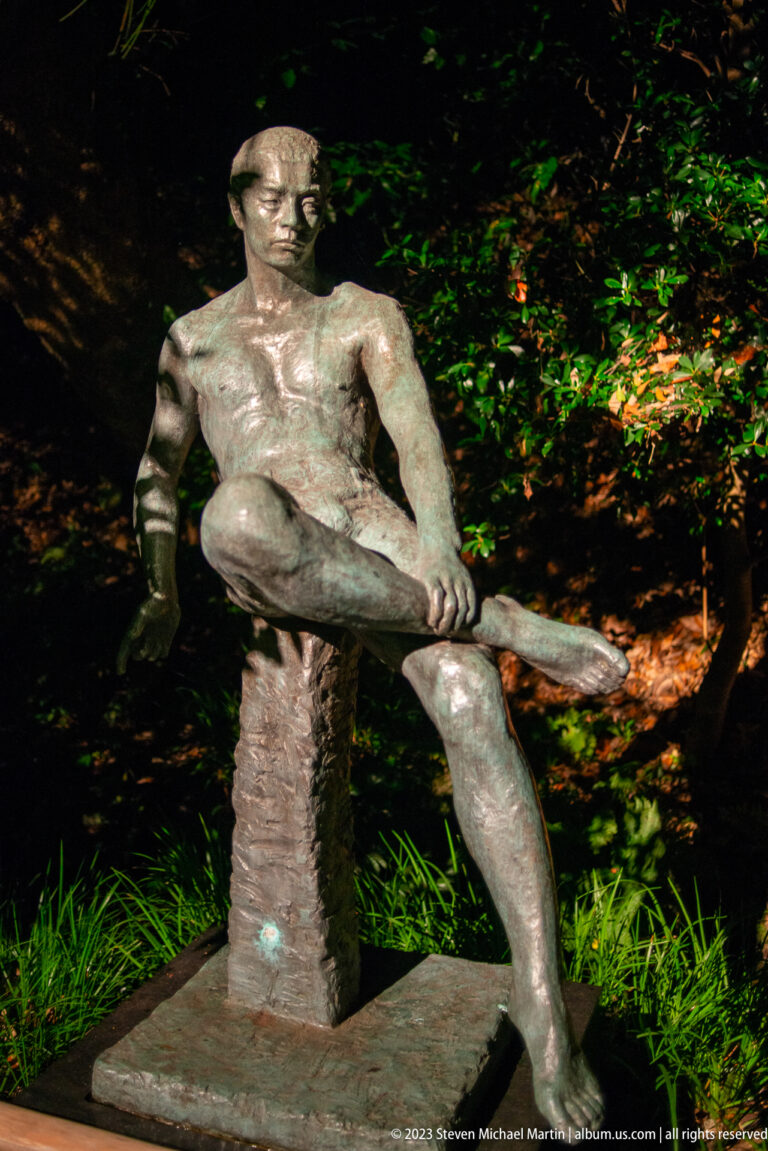Bronze sculpture of a seated man with one leg raised, illuminated at night in a garden.