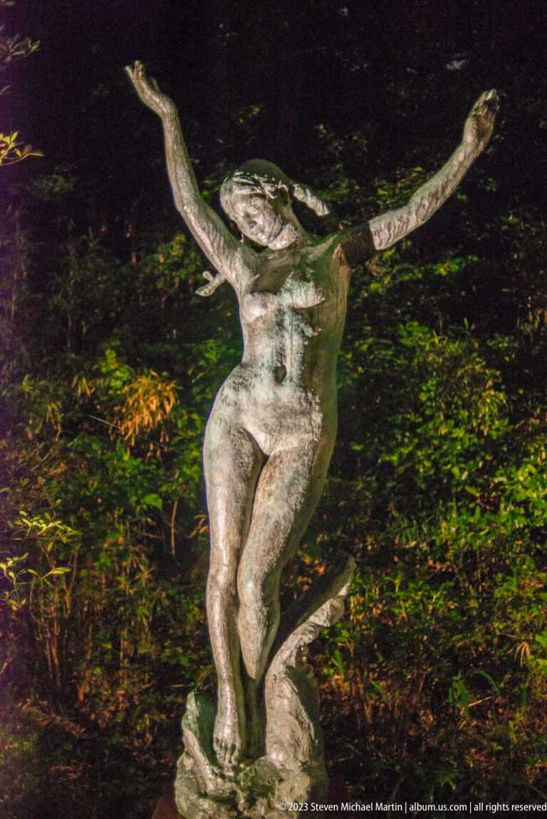 Bronze female figure with both arms raised, illuminated against dark garden foliage.