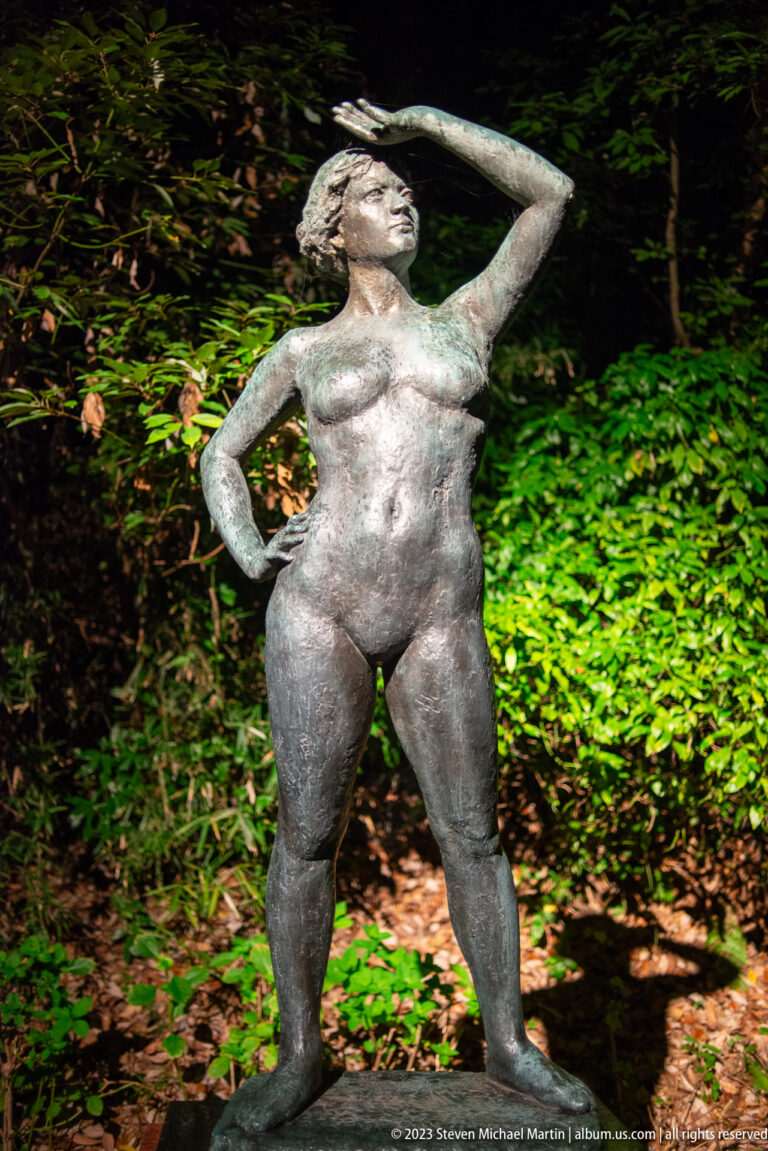 Standing bronze woman with one hand at her forehead and the other on her hip.
